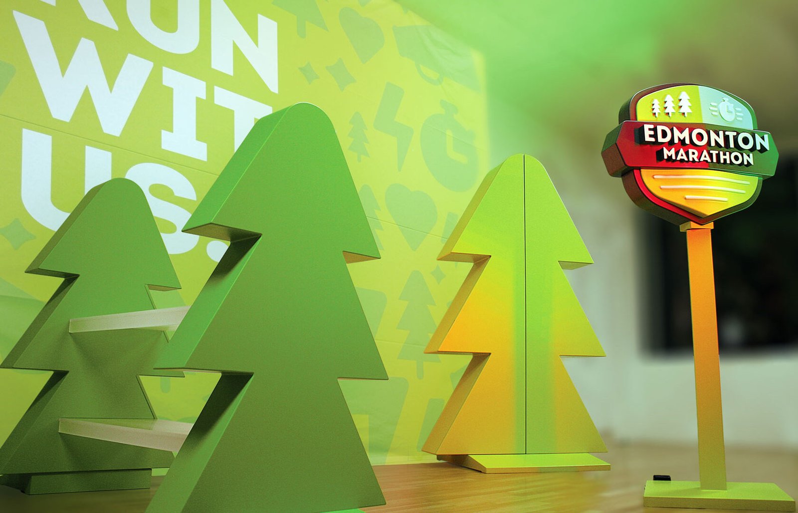Trade_Show_Booth Custom trade show booth fabrication for the Edmonton Marathon, comprised of a yellow tree shaped display cabinet, a green tree shaped display with acrylic shelving and a colourful backdrop reading "Run With Us".