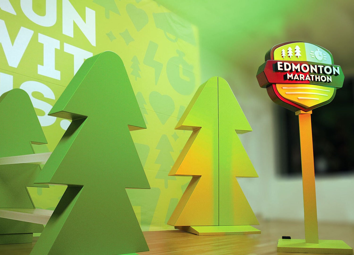 Trade_Show_Booth Custom trade show booth fabrication for the Edmonton Marathon, comprised of a yellow tree shaped display cabinet, a green tree shaped display with acrylic shelving and a colourful backdrop reading "Run With Us".