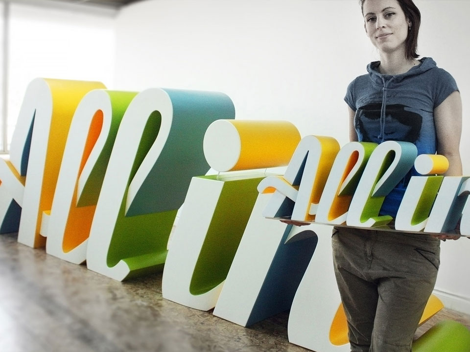 Woman holding a 3 dimensional logo prop reading All In, posing in front of a giant 18 feet long giant logo sign.