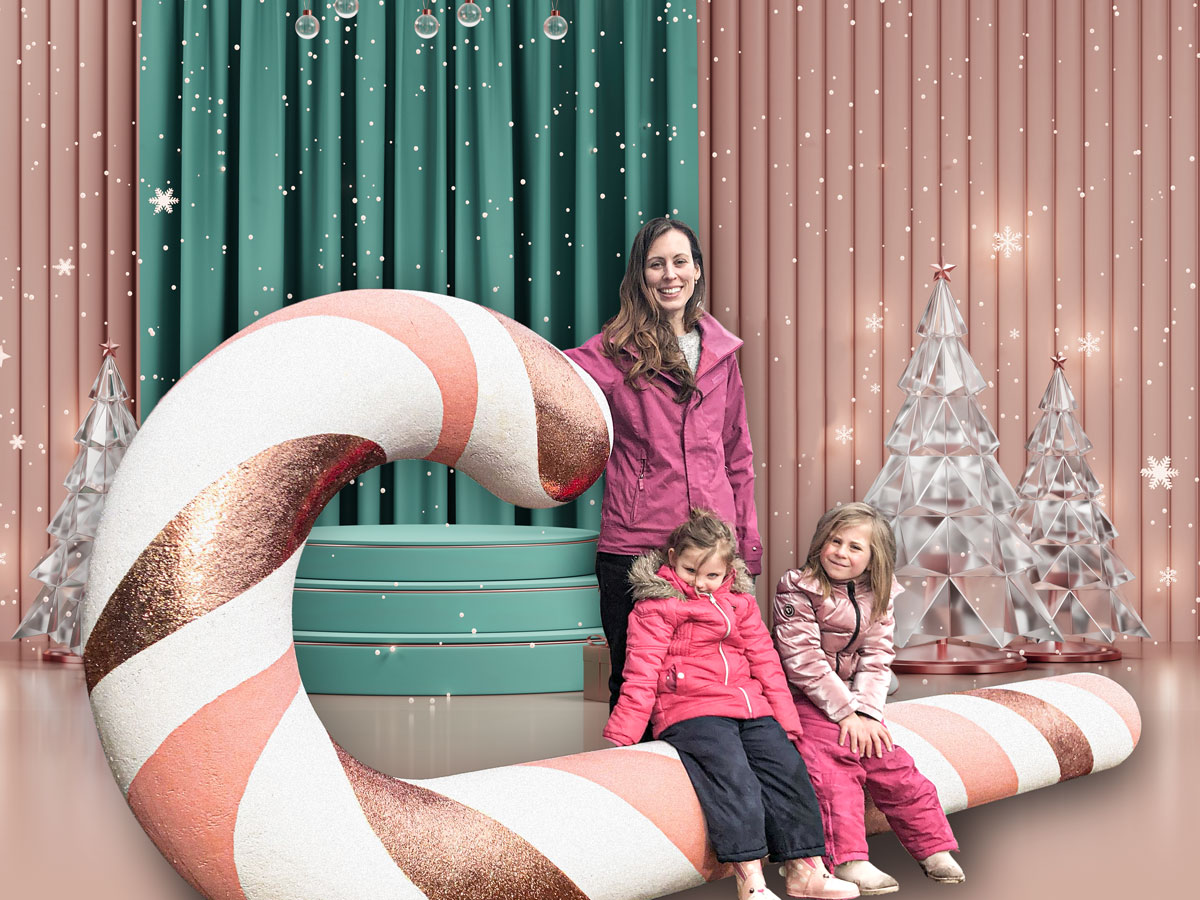 A woman and 2 kids sitting on a giant 12 foot long candy cane prop.