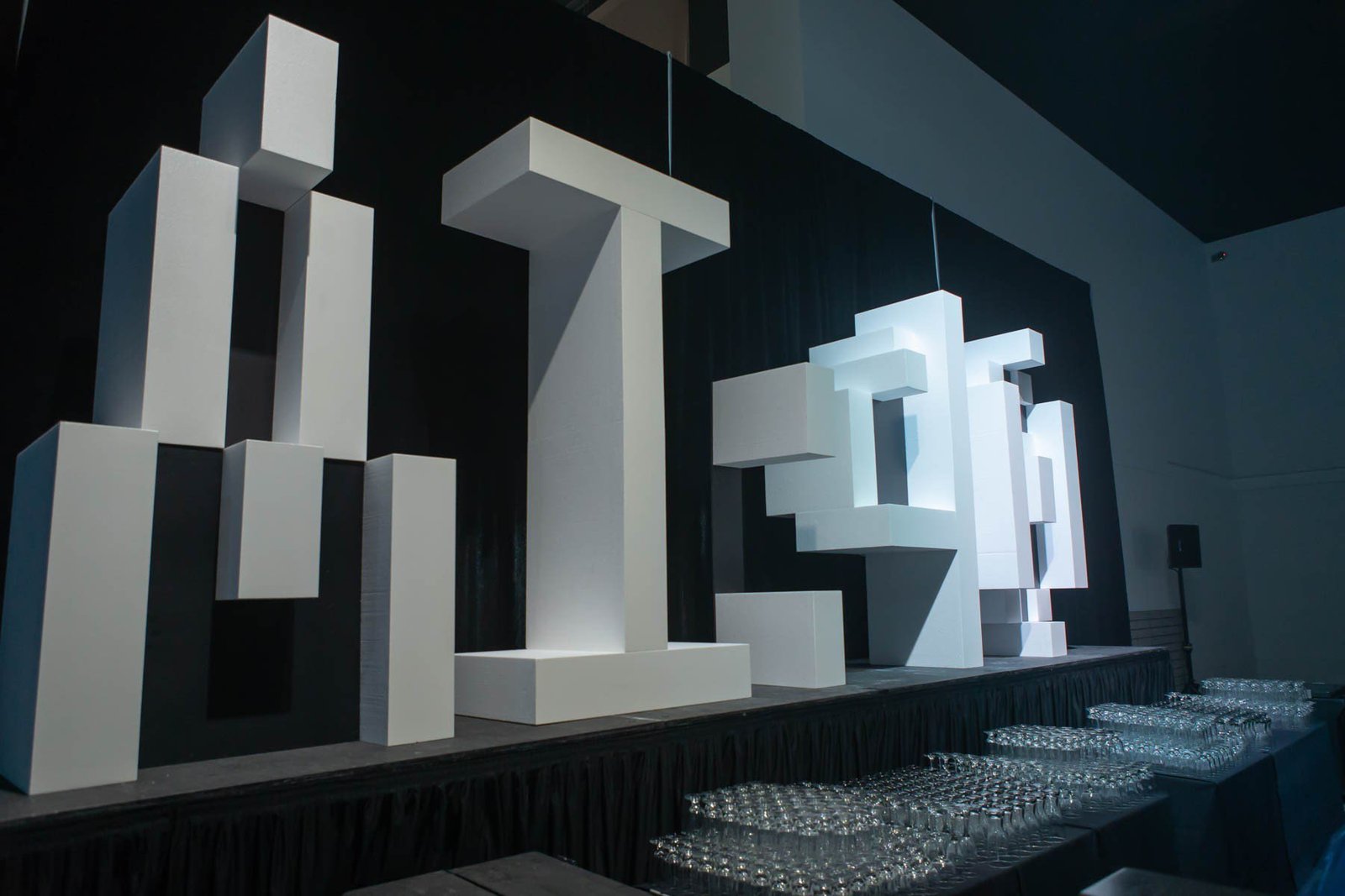 1588637313634_134-studio-e-4X6-EDJ_8223 Custom stage props, abstract white geometric block sculptures arranged on a dark stage backdrop, with rows of glassware lining the front edge of a table/venue floor.