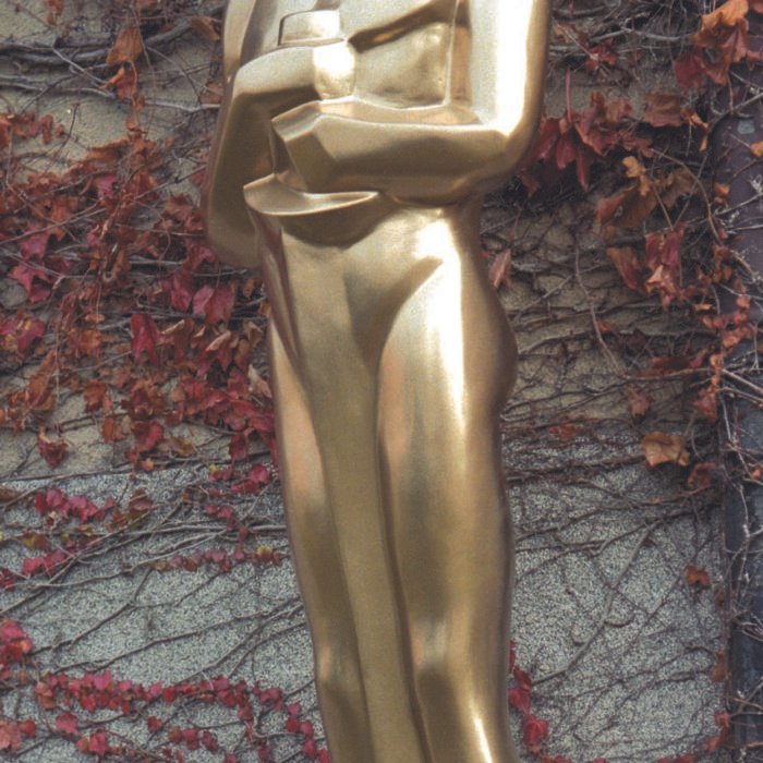 Oskar - The Bees Knees Design Gold Oscar statue standing outdoors on a pedestal, with ivy-covered wall in the background. Styrofoam sculpture