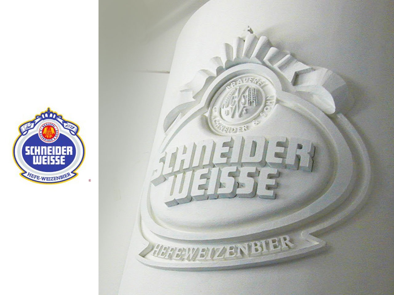 White embossed Schneider Weisse logo on a wall, with a small blue-and-gold Schneider Weisse crest to the left and the words 'HEFEWEIZENBIER' below.