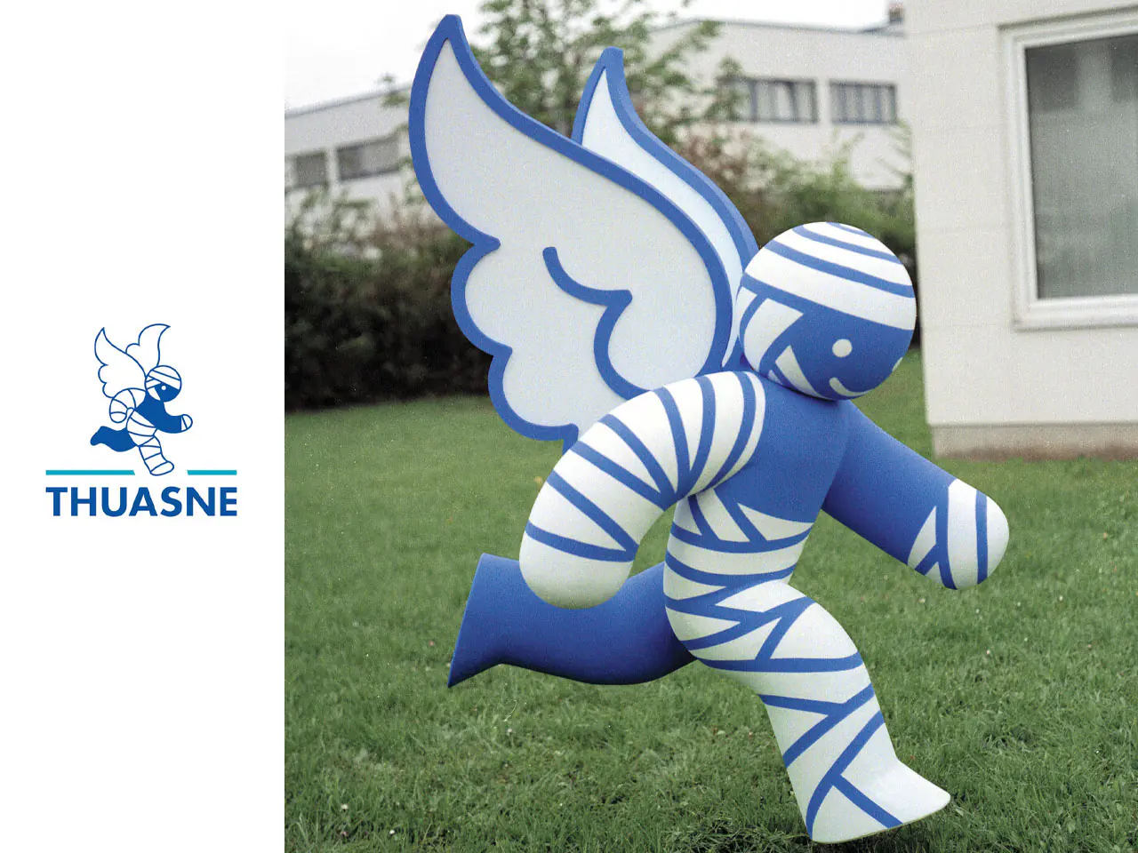 3D_Caroussel_Thuasne - The Bees Knees Design Thuasne logo with a blue winged mascot on the left and a large blue-and-white striped inflatable winged figure running on grass to the right. 2d to 3d logo