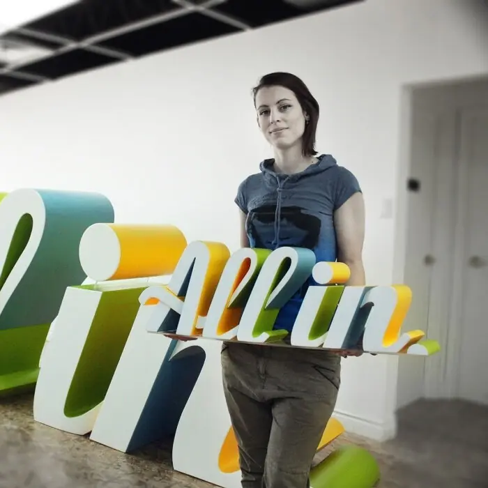 Woman in casual clothing holding a large colorful 3D typography sculpture in a bright studio, posing for the camera.