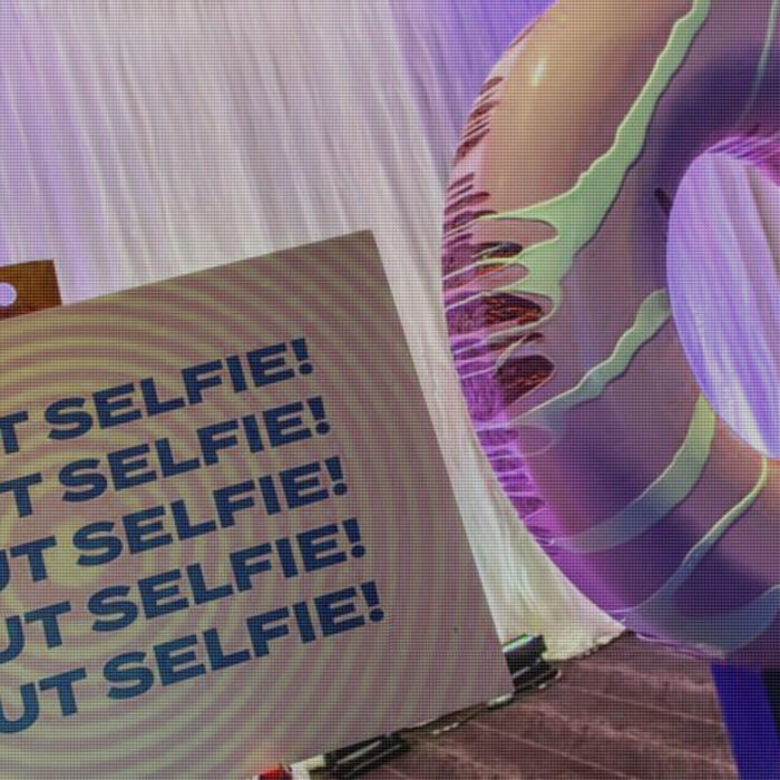 A beige display box printed with 'DONUT SELFIE!' repeated, placed next to a large pink frosted donut on a purple-lit background.