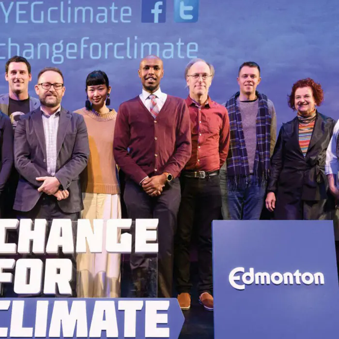 Ten diverse adults pose on a stage for a climate-change event, with a large 'CHANGE FOR CLIMATE' sign and an Edmonton banner visible.