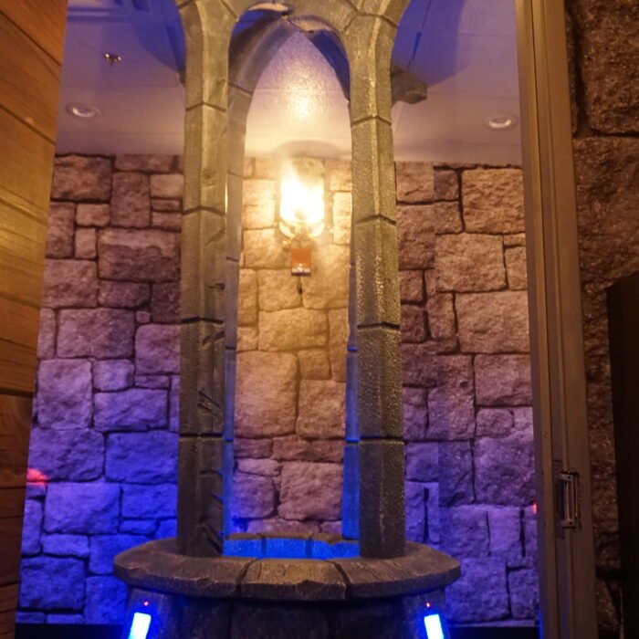 DSC03488 - The Bees Knees Design Stone fountain with four vertical pillars forming an arch over a circular basin, set against a rough stone wall and warm overhead light.