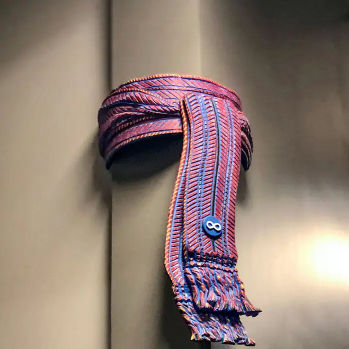 Exhibits_Metis_Sash - The Bees Knees Design Blue and red striped scarf with fringed ends looped over a metallic wall panel.