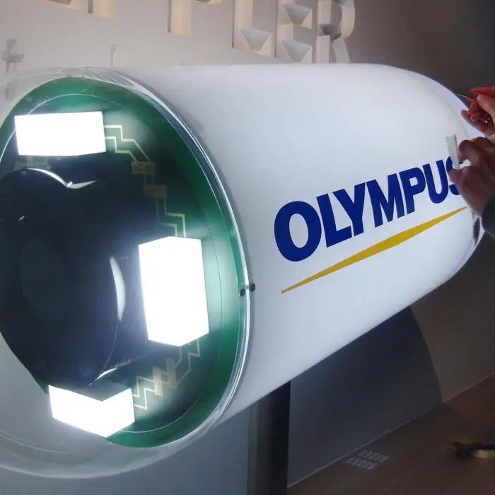 Exhibits_Olympus - The Bees Knees Design Technician working on a large Olympus medical device, using red-handled tools near a white cylindrical unit with the blue 'OLYMPUS' logo visible on the side.