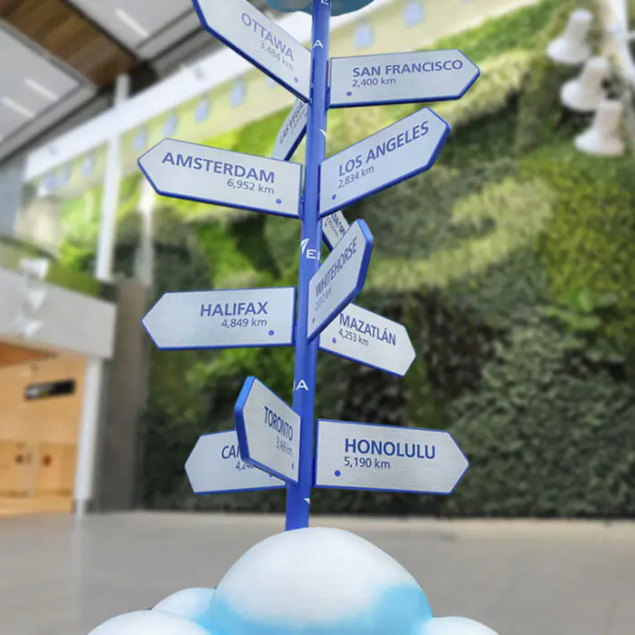 Blue directional signpost in a modern atrium with arrows pointing to cities like Amsterdam, Los Angeles, San Francisco and more.