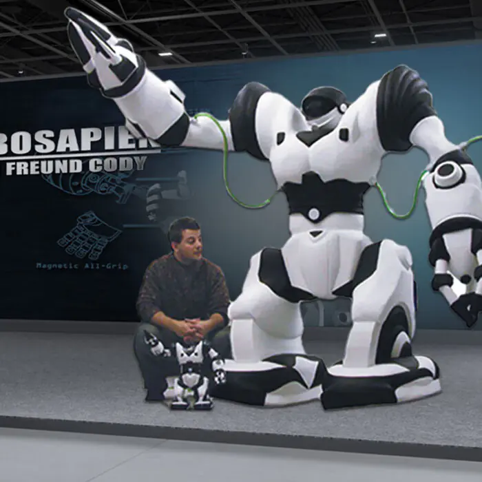Life-sized white and black Robosapien robot with outstretched arms on display, connected by green cables, beside a man and two small robots.