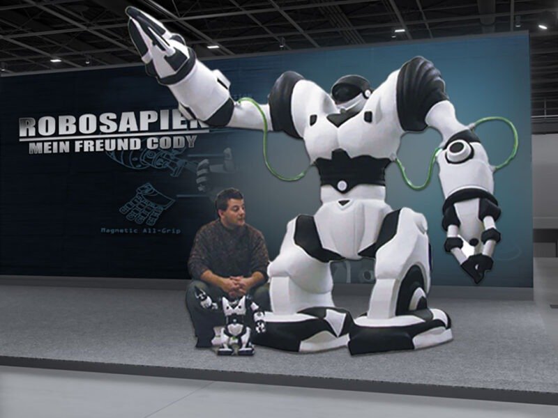 Product_Replicas_RoboSapien_2 Life-sized white and black Robosapien robot experiential photo booth with outstretched arms on display, connected by green cables, beside a man and two small robots.