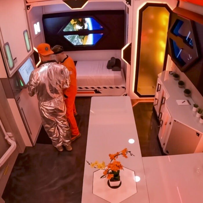 Screenshot - The Bees Knees Design Two astronauts in orange and silver suits at a control panel inside a white space habitat, Earth visible through a window beyond them.