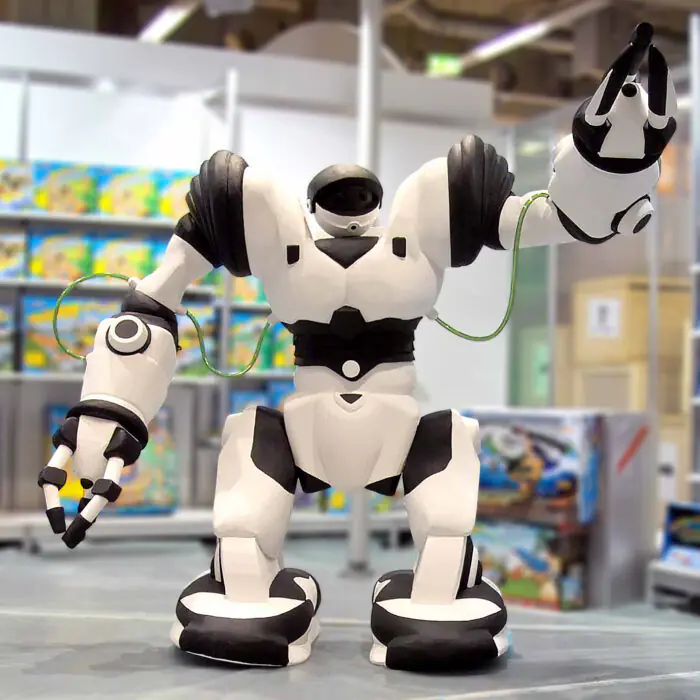 Large white humanoid robot with black accents, arms raised, cables looping from its arms in a toy store display.
