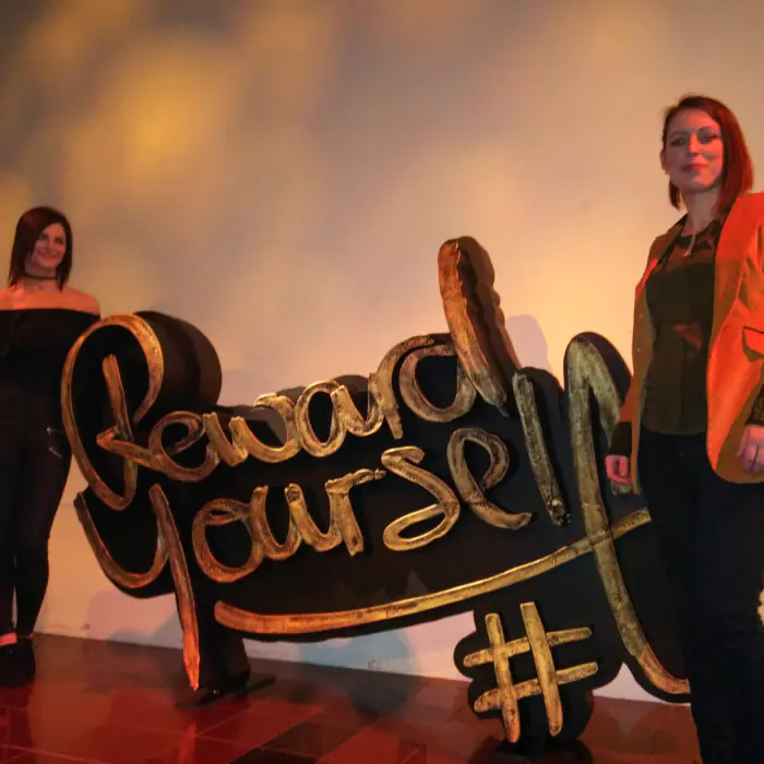 Two women pose on a stage beside a large gold-script sign, with warm lighting in the background.