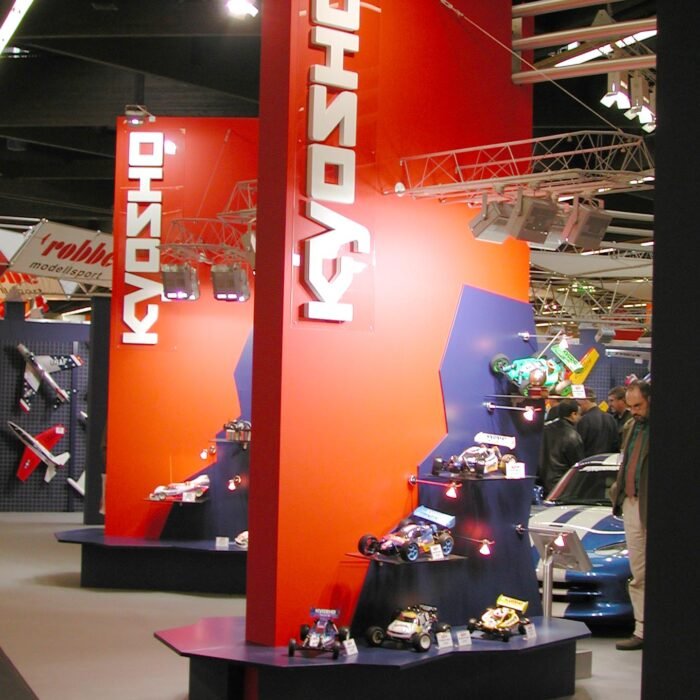 Exhibition booth with tall red pillars and white vertical 'KYOSHO' text, displaying model cars on black platforms under bright lighting.
