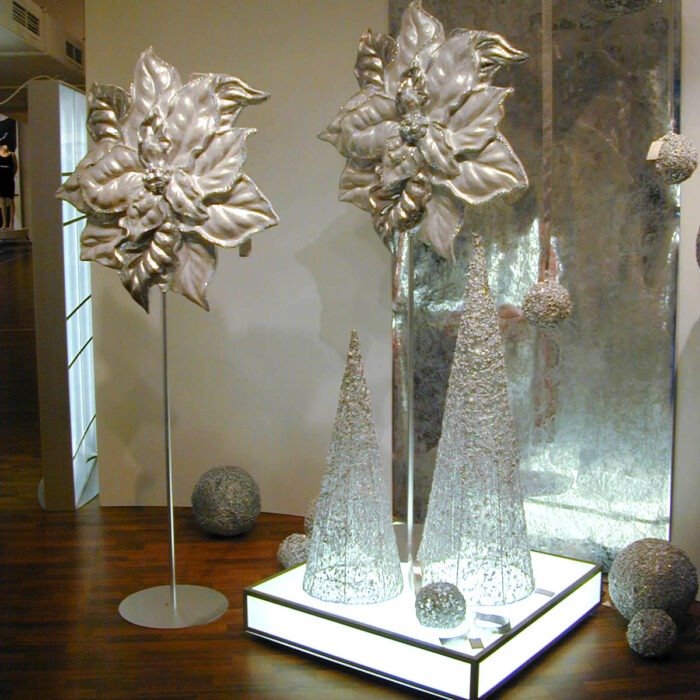 Silver metallic flower sculptures on slender stands displayed on an illuminated square base, with sculpted cone trees and textured spheres nearby in a gallery.