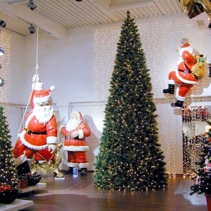 Indoor Christmas display with a tall, illuminated Christmas tree and oversized Santa figures, including a hanging Santa rope-pulled prop, in a showroom with string lights on white walls. Mall decor