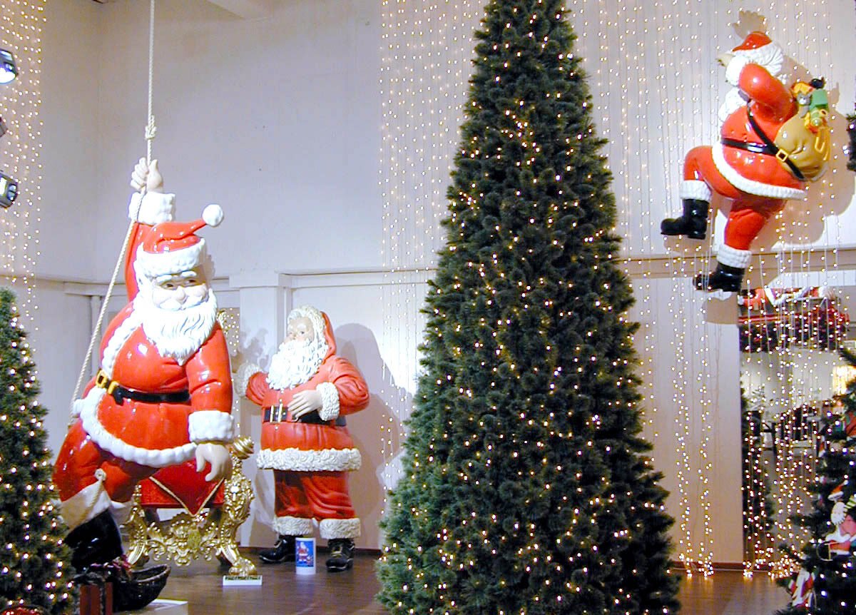 deco weihnacht4 Indoor christmas retail display with a tall, illuminated Christmas tree and oversized Santa figures, including a hanging Santa rope-pulled prop, in a showroom with string lights on white walls.